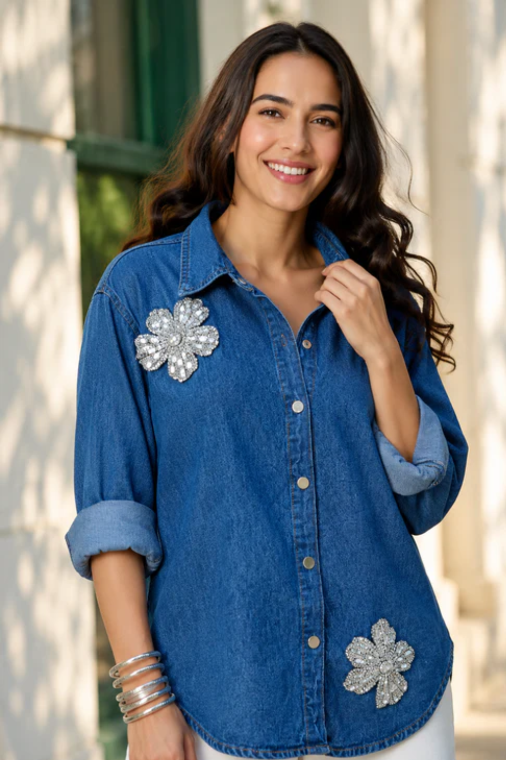 A woman wearing a blue denim Denim Shirt for women with two large silver rhinestone flower appliqués, one on the chest and one near the lower front hem, posed outdoors against a softly blurred light-colored building background.