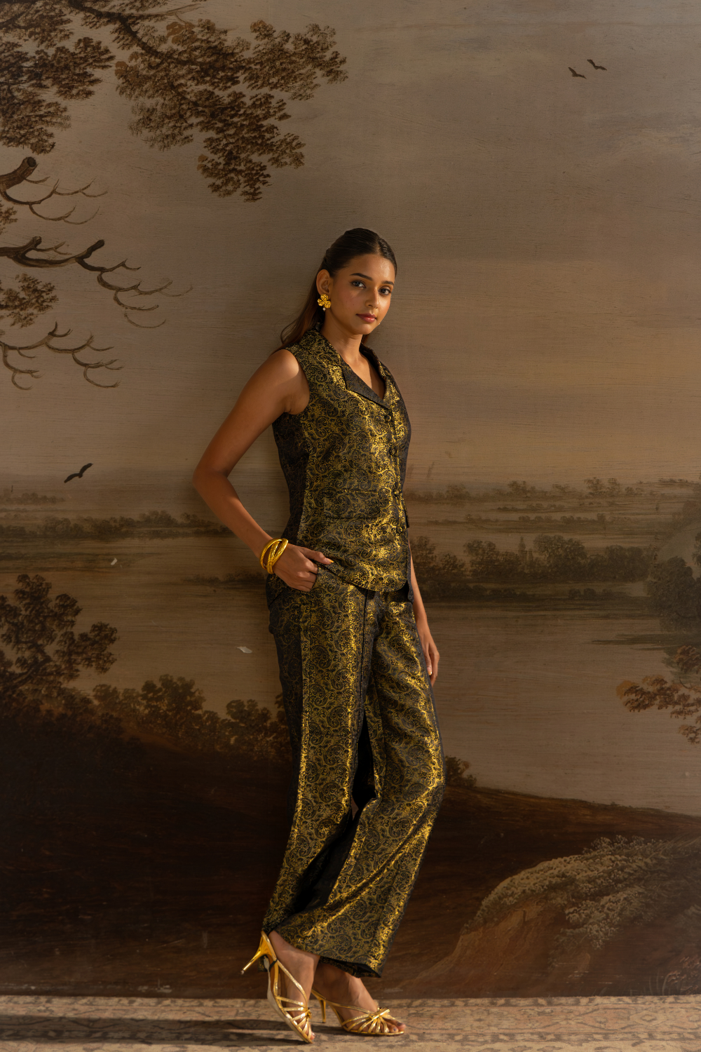 A woman models a sleeveless black and gold patterned vest and matching wide-leg trousers in brocade fabric , styled with gold jewelry and heels, set against a scenic landscape mural.