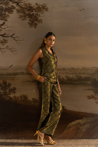 A woman models a sleeveless black and gold patterned vest and matching wide-leg trousers in brocade fabric , styled with gold jewelry and heels, set against a scenic landscape mural.
