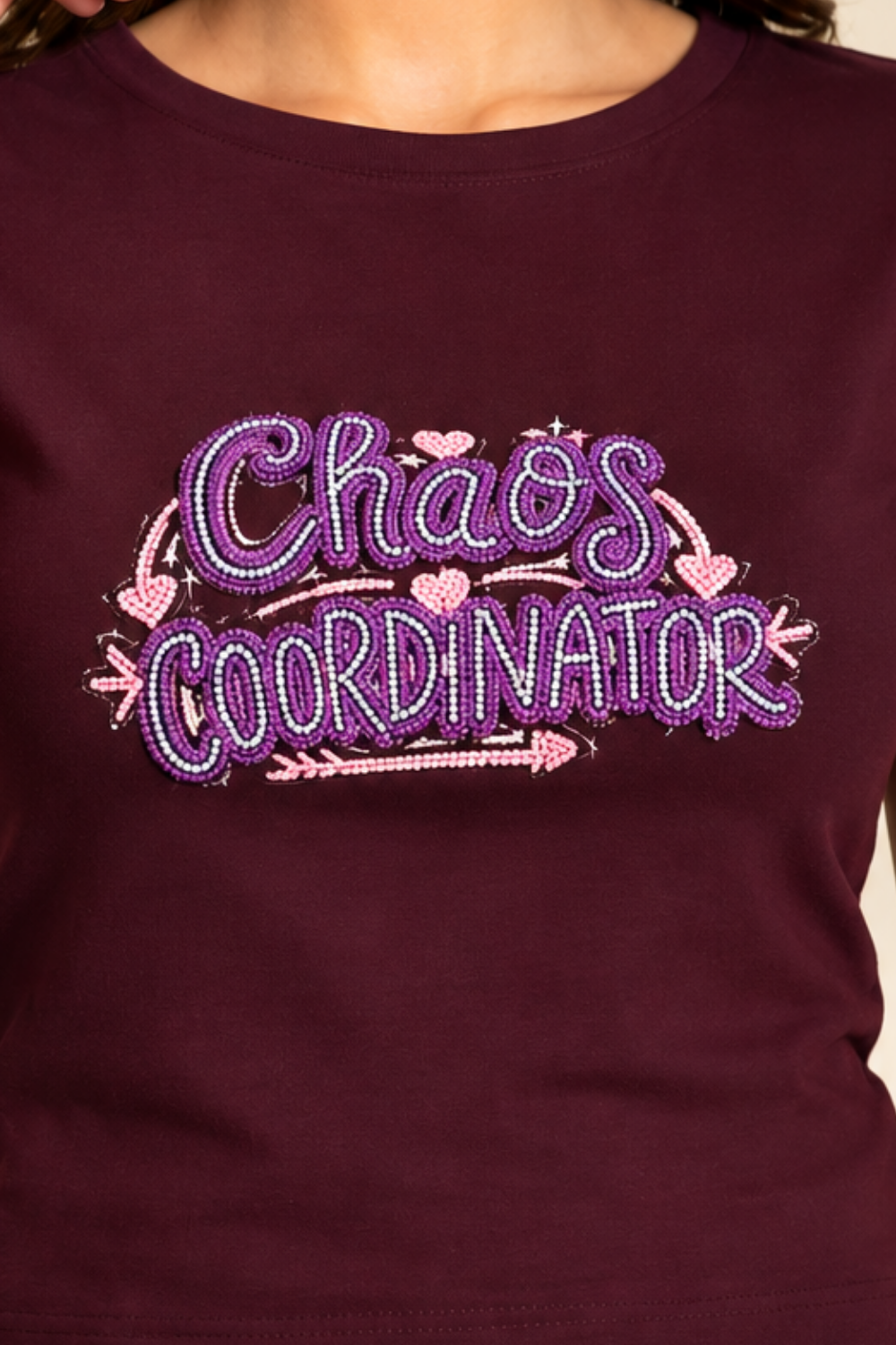 A close-up of a person wearing a dark burgundy t-shirt with a glittery purple and pink rhinestone graphic that reads 'Chaos Coordinator' with hearts and arrows.