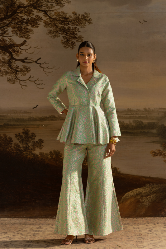 A woman stands in front of a scenic landscape backdrop wearing a light sage green two-piece suit with a subtle floral pattern, featuring a peplum-style blazer and matching wide-leg pants.