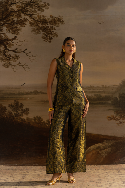 A woman models a sleeveless button-front vest and matching wide-leg trousers in a metallic gold and black damask pattern, posed against a classical landscape painting backdrop.