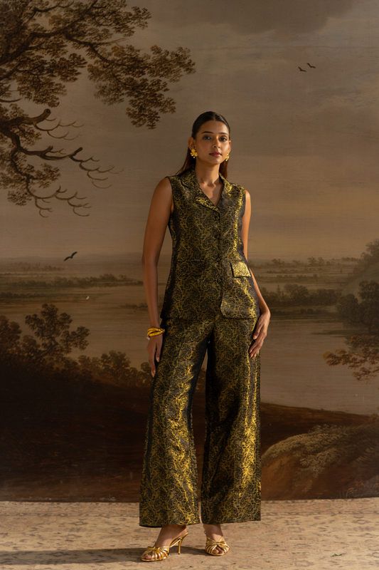 A woman models a sleeveless button-front vest and matching wide-leg trousers in a metallic gold and black damask pattern, posed against a classical landscape painting backdrop.