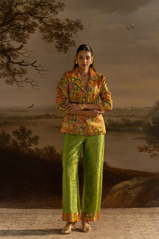 A woman stands in front of a painted landscape backdrop wearing a colorful patterned long-sleeve blazer and bright green wide-leg trousers with orange border detailing at the hem.