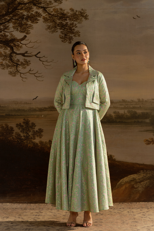 A woman stands in front of a large classical landscape painting, wearing a light sage green brocade dress with a matching cropped jacket. The dress features a delicate floral pattern and a full skirt, paired with a structured jacket that has wide lapels and flap pockets.