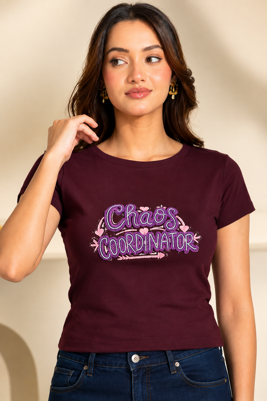 A woman wearing a maroon short-sleeve t-shirt with a purple and pink graphic that reads 'Chao's Coordinator' with hearts and arrows, paired with dark blue jeans against a warm beige background.