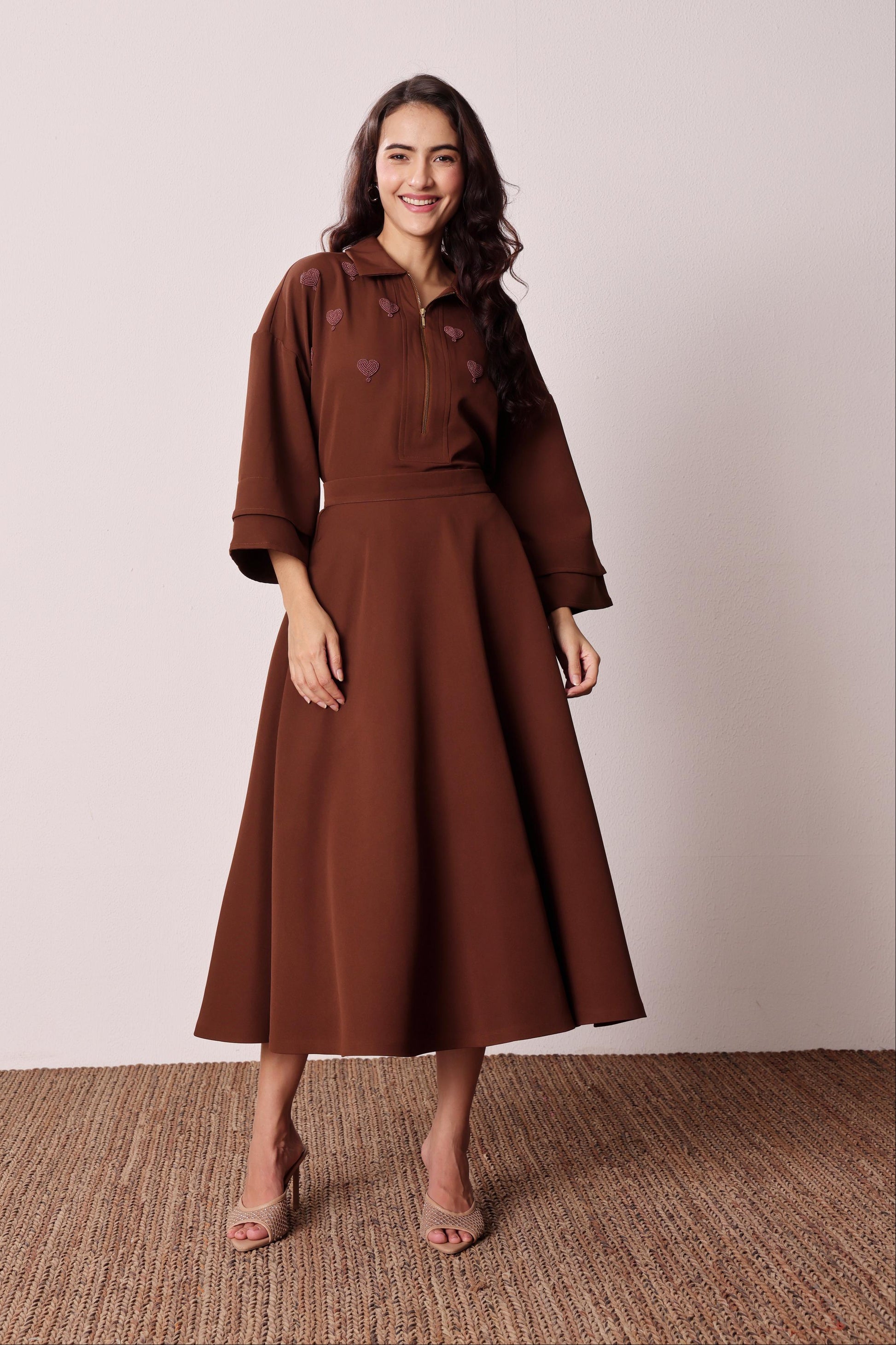A woman modeling a brown midi dress with three-quarter sleeves, a collared neckline, and embroidered heart details on the chest, standing against a neutral beige wall and textured brown floor.