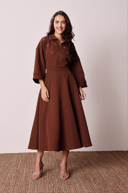 A woman modeling a brown midi dress with three-quarter sleeves, a collared neckline, and embroidered heart details on the chest, standing against a neutral beige wall and textured brown floor.